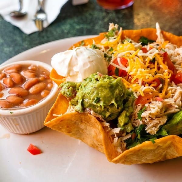 A taco salad in a crispy shell with chicken, guacamole, sour cream, cheese, and a side of beans.